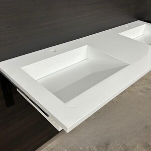 May include: A bright white, rectangular bathroom sink with two integrated basins. The sink has a clean, modern design with a smooth, glossy finish. The sink is mounted on a dark brown surface.