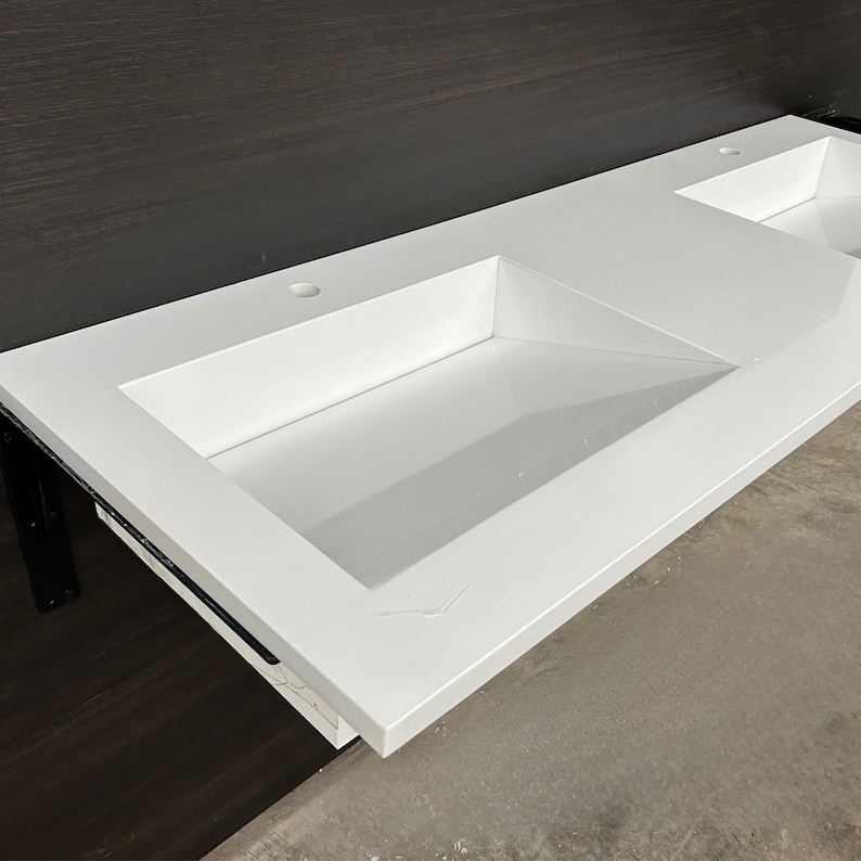 May include: A bright white, rectangular bathroom sink with two integrated basins. The sink has a clean, modern design with a smooth, glossy finish. The sink is mounted on a dark brown surface.
