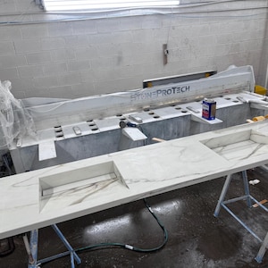 May include: A large, white countertop with a marble-like pattern, featuring two rectangular cutouts. The countertop is supported by metal stands. A large machine with the text "STONE PROTECH" is in the background, along with a control panel.