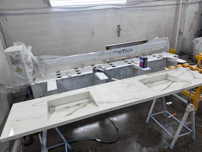 May include: A large, white countertop with a marble-like pattern, featuring two rectangular cutouts. The countertop is supported by metal stands. A large machine with the text "STONE PROTECH" is in the background, along with a control panel.