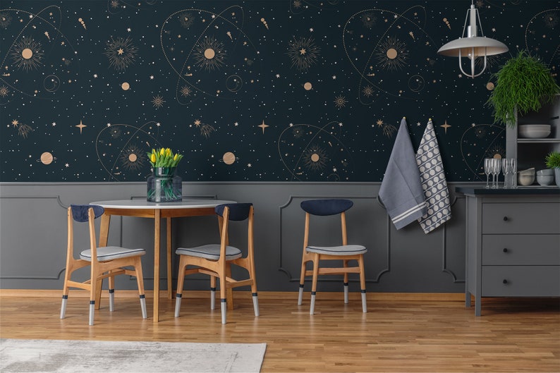 Op de afbeelding: Een eetkamer met donkerblauwe behang met een gouden hemels patroon. De behang heeft sterren, planeten en sterrenbeelden. Er is een houten tafel met vier stoelen, waarvan er twee zichtbaar zijn op de afbeelding. De stoelen hebben blauwe kussens en houten poten. Aan de rechterkant van de afbeelding is een grijze kast met drie lades.