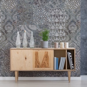 Puede incluir: Un armario de madera con tres estantes y dos puertas. El armario es de color marrón claro con un acabado de madera natural. El armario está decorado con tres botellas de vidrio transparente y una pequeña planta en maceta. El armario está delante de una pared con un papel tapiz estampado en gris y blanco.