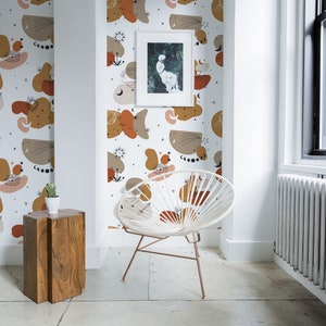 Puede incluir: Una silla blanca con un diseño tejido se encuentra en una habitación con paredes blancas y un papel tapiz estampado con formas abstractas en tonos marrones, naranjas y blancos. El papel tapiz tiene un tema celestial con estrellas, lunas y ojos.