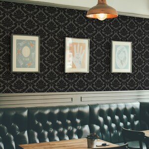 Op de afbeelding: Een zwart-wit damast behangpatroon met drie ingelijste foto's die aan de muur hangen. De foto's zijn van een hand met een anker-tatoeage, een groep mensen en een gezicht. Op de voorgrond is een donkergroene leren bank met een houten tafel en stoelen.