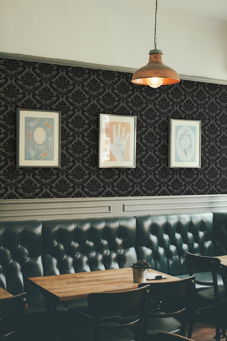 Op de afbeelding: Een zwart-wit damast behangpatroon met drie ingelijste foto's die aan de muur hangen. De foto's zijn van een hand met een anker-tatoeage, een groep mensen en een gezicht. Op de voorgrond is een donkergroene leren bank met een houten tafel en stoelen.
