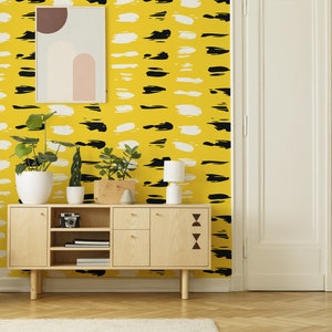 Könnte beinhalten: Ein Holzschränkchen mit drei Regalen und zwei Schubladen, hinter dem sich eine Tapete mit abstrakten Mustern in Schwarz und Weiß befindet. Das Schränkchen ist mit zwei Topfpflanzen und einer Lampe dekoriert.