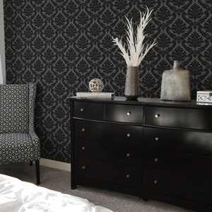 Op de afbeelding: Een zwarte commode met zes laden en zilveren knoppen. De commode is versierd met een vaas met witte veren, een zilveren vaas, een decoratieve bol en een zilveren doos. De commode staat voor een zwarte damastbehang.