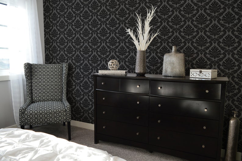 Op de afbeelding: Een zwarte commode met zes laden en zilveren knoppen. De commode is versierd met een vaas met witte veren, een zilveren vaas, een decoratieve bol en een zilveren doos. De commode staat voor een zwarte damastbehang.