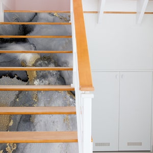 Puede incluir: Un conjunto de escaleras de madera con un dise&ntilde;o abstracto gris, negro y dorado en cada escal&oacute;n. Las escaleras est&aacute;n en una casa con paredes blancas y una puerta blanca.
