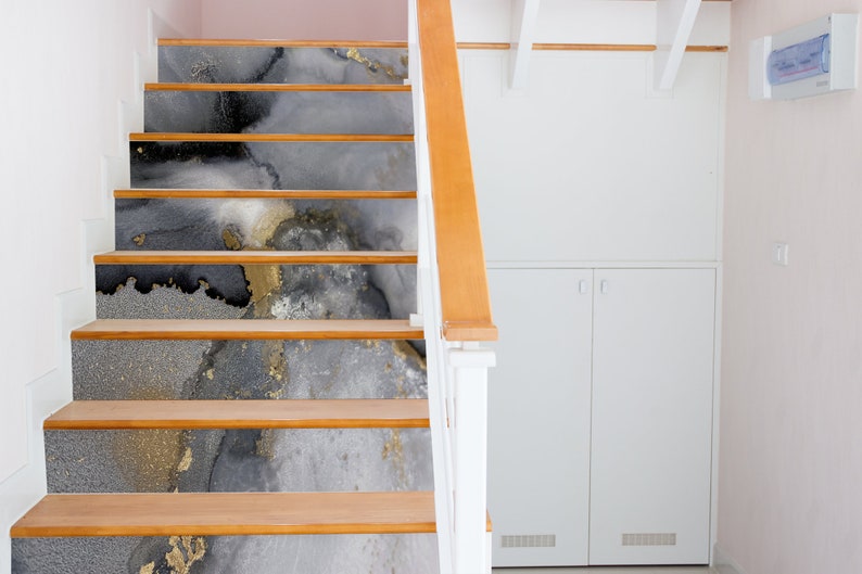 Puede incluir: Un conjunto de escaleras de madera con un dise&ntilde;o abstracto gris, negro y dorado en cada escal&oacute;n. Las escaleras est&aacute;n en una casa con paredes blancas y una puerta blanca.