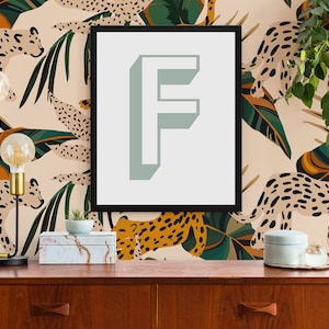 Peut inclure: Une impression encadr&eacute;e noire avec une lettre "F" vert clair sur fond blanc. L'impression est accroch&eacute;e &agrave; un mur avec un fond beige orn&eacute; d'un motif r&eacute;p&eacute;titif de l&eacute;opards stylis&eacute;s et de feuilles tropicales dans des tons de vert, orange et marron.