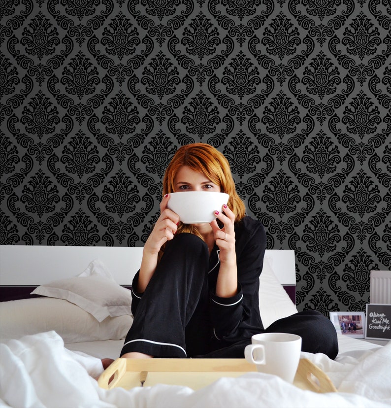 Op de afbeelding: Een vrouw in een zwarte pyjama zit op een bed met een witte kom voor haar gezicht. Het bed heeft een wit dekbed en een wit kussen. De muur achter haar is bedekt met een zwart-witte damastbehang.