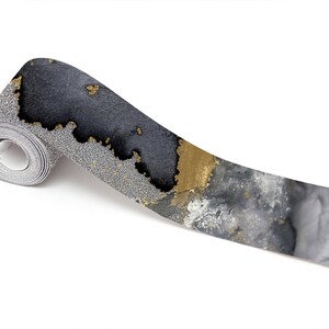 Puede incluir: Un rollo de cinta washi decorativa con un dise&ntilde;o abstracto en negro, dorado y plateado. La cinta est&aacute; enrollada y el extremo est&aacute; desenrollado, mostrando el dise&ntilde;o.