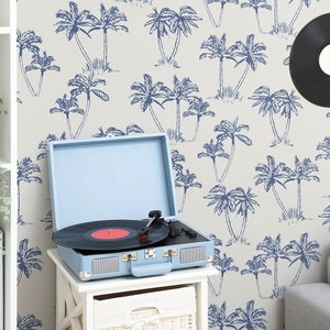 May include: A light blue record player with a black vinyl record on the turntable. The record player is sitting on a white table with a wicker basket underneath. The background is a light gray wall with a blue palm tree pattern.