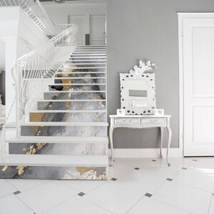 Puede incluir: Una escalera blanca con un dise&ntilde;o abstracto gris y dorado en cada escal&oacute;n. La escalera est&aacute; en una casa moderna con paredes blancas y una barandilla blanca.