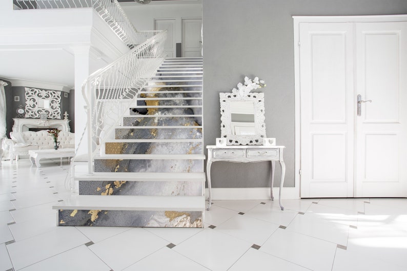 Puede incluir: Una escalera blanca con un dise&ntilde;o abstracto gris y dorado en cada escal&oacute;n. La escalera est&aacute; en una casa moderna con paredes blancas y una barandilla blanca.