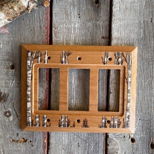 May include: A wooden light switch cover with a birch tree design. The cover has three switch openings and is made of natural wood.