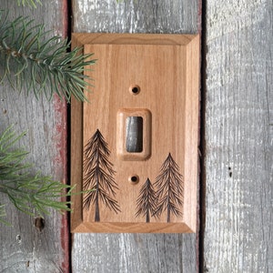 May include: A light brown wooden light switch cover with a laser-etched design of three pine trees. The switch cover is mounted on a weathered wooden wall.