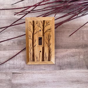 May include: A wooden light switch cover with a woodburned design of trees. The cover is a light brown color and the trees are a darker brown.