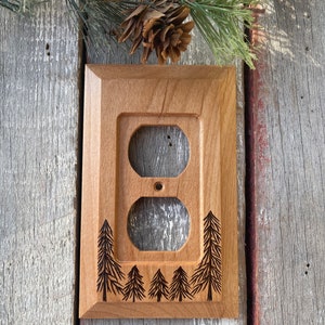 May include: A wooden outlet cover with a natural finish. The cover features a rectangular frame with two outlet holes and a single screw hole. The design includes etched pine trees along the bottom and sides. A pine branch and pine cone are in the background.