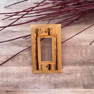 May include: A wooden light switch cover with a burnt wood design of trees. The cover is a light brown color and has a rectangular shape.
