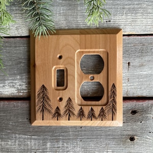 May include: A wooden light switch and outlet cover with a laser-etched design of a forest of pine trees. The cover is made of natural wood and has a light brown color.