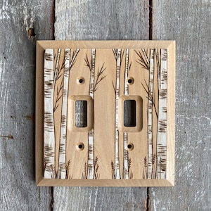 May include: A wooden light switch cover with a birch tree design. The cover is light brown with a darker brown border. The birch trees are white with black lines.