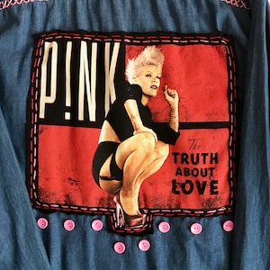 Könnte beinhalten: Eine Jeansjacke mit einem rosa und schwarzen Patch, auf dem eine Sängerin mit dem Text "P!NK The Truth About Love" abgebildet ist. Der Patch ist von rosa Nähten umgeben. Die Jacke hat rosa Knöpfe.