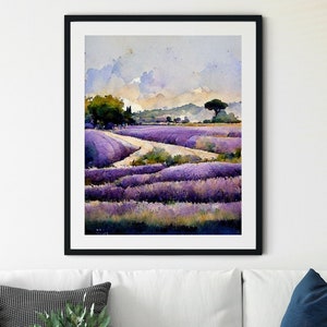 May include: A framed watercolor painting of a lavender field with a dirt path leading through it. The painting is set against a white wall in a living room setting with a white couch, a throw blanket, and a side table.