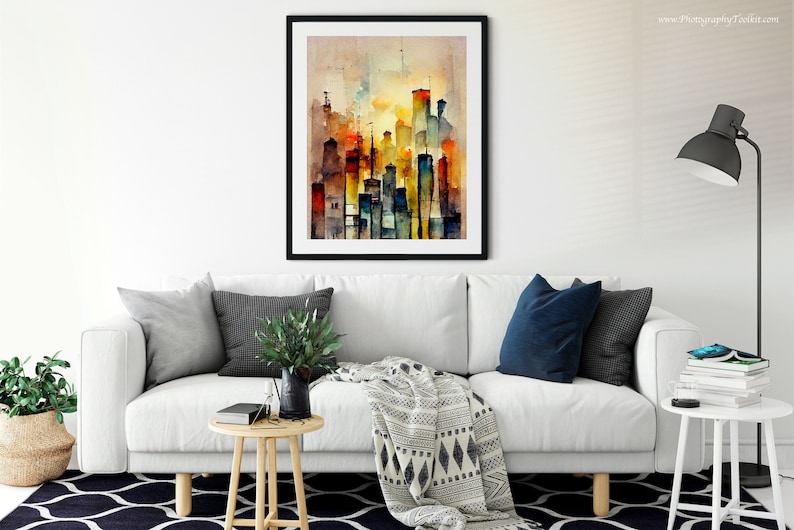 May include: A watercolor painting of a city skyline in shades of orange, yellow, blue, and gray. The painting is framed in a black frame and is hanging on a white wall above a white couch.