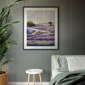 May include: A framed watercolor painting of a lavender field with a dirt road leading through it. The painting is in a black frame and is hanging on a gray wall. There is a white side table with a magazine on it in the foreground.