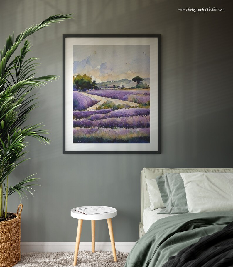 May include: A framed watercolor painting of a lavender field with a dirt road leading through it. The painting is in a black frame and is hanging on a gray wall. There is a white side table with a magazine on it in the foreground.