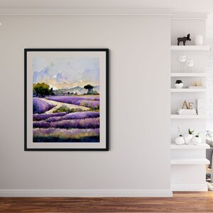 May include: A framed watercolor painting of a lavender field with a winding path leading to a small village in the distance. The sky is a soft blue with fluffy white clouds. The painting is in a black frame and is hanging on a white wall.