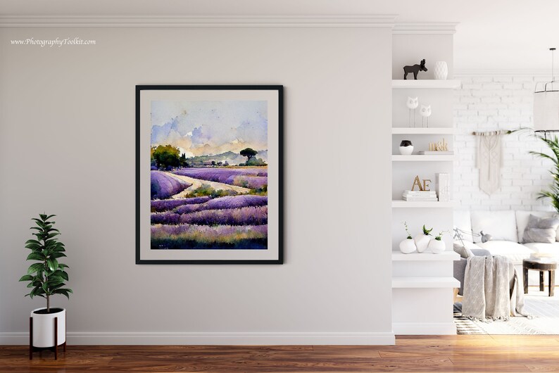May include: A framed watercolor painting of a lavender field with a winding path leading to a small village in the distance. The sky is a soft blue with fluffy white clouds. The painting is in a black frame and is hanging on a white wall.