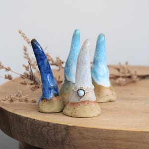 Könnte beinhalten: Vier Keramik-Ringhalter in Blau-, Weiß- und Beigetönen. Jeder Halter hat eine einzigartige Form und einen Farbverlauf, mit einem Ring auf einem. Die Halter sind auf einer Holzoberfläche mit getrockneten Pflanzen angeordnet.