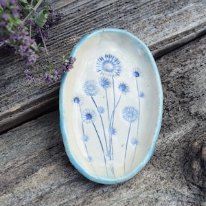 Könnte beinhalten: Eine ovale Keramikschale mit cremefarbener Basis und hellblauem Rand. Das Innere zeigt ein handgemaltes blaues Blumenmuster, das mehrere zarte Wildblumen mit dünnen Stielen und kreisförmigen, strahlenden Blütenblättern darstellt.