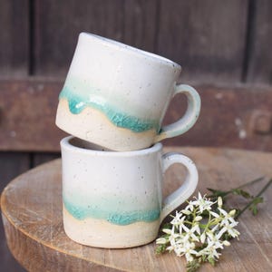 Op de afbeelding: Twee gestapelde keramische mokken met een gespikkelde witte afwerking en een turquoise blauw druipglazuurpatroon bij de basis. Elke mok heeft een stevig handvat en een cilindrische vorm, met een duidelijk contrast tussen het lichte gespikkelde bovendeel en het levendige blauw geglazuurde onderste gedeelte.