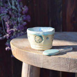 Könnte beinhalten: Eine handgefertigte Keramiktasse mit hellgelbem Boden und türkisfarbenem Rand und Akzenten. Die Tasse hat geprägte Blumendesigns. Ein kleiner, passender Löffel liegt daneben auf einem Holztisch. Violette Blumen sind im Hintergrund.
