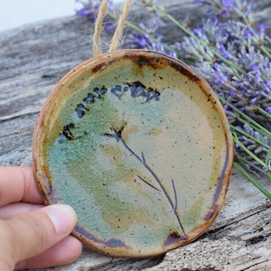 Op de afbeelding: Een ronde keramische ornament met een bloemenontwerp. Het ornament heeft een beige en groene achtergrond met een donkerbruine rand. Een takje lavendel is op het ornament geschilderd. Er is een touwlus bevestigd om op te hangen.