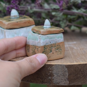 Könnte beinhalten: Zwei kleine, handgefertigte Keramikgläser mit Deckel. Die Gläser sind quadratisch mit hellbraunem Boden, einem türkisfarbenen und weißen Oberteil und einem braunen Deckel. Der untere Teil hat ein Blumendesign. Ein Glas wird von einer Hand gehalten.