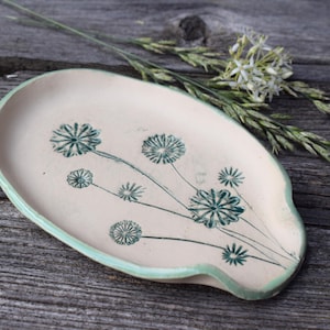 Può includere: Un poggia cucchiaio in ceramica di forma ovale con base color crema e bordo verde chiaro. La superficie presenta un'illustrazione botanica verde scuro di diversi fiori simili a denti di leone con steli sottili e teste circolari al centro.