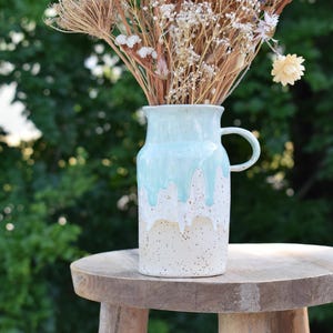Könnte beinhalten: Eine Keramikvase mit hellblauer Glasur, die über eine gesprenkelte weiß-beige Basis tropft. Die Vase hat einen Henkel und ist mit getrockneten Blumen und Gräsern gefüllt. Sie steht auf einem kleinen Holzschemel.