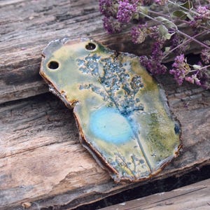 May include: A handcrafted ceramic pendant with an irregular form. The pendant displays a mix of green, yellow, and blue tones, with a central blue area. Two holes are at the top. It rests on a weathered wooden surface, accompanied by purple flowers.