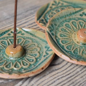 Könnte beinhalten: Drei runde Räucherstäbchenhalter aus Keramik mit grüner Craquelé-Glasur und einem erhöhten beigen Knopf in der Mitte, der ein Loch für ein Räucherstäbchen aufweist. Die Teller haben ein geprägtes Blumenmuster und sichtbare Terrakotta-Ränder.