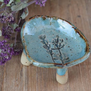 Könnte beinhalten: Eine handgefertigte Keramikschale mit hellblauer Glasur und floralem Design. Die Schale wird von drei kleinen Beinen getragen und hat einen braunen Rand. Im Hintergrund sind lila Blumen zu sehen. Die Schale hat einen Durchmesser von etwa 10 cm.