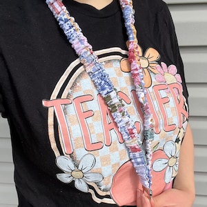 May include: A black t-shirt with the word "TEACHER" in a retro design. The shirt features a floral and checkerboard pattern. A floral patterned lanyard with a metal clasp is around the neck.