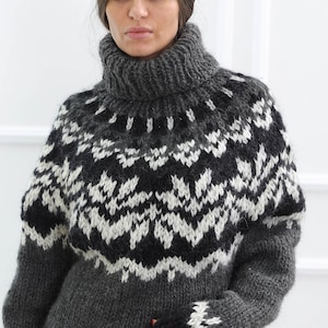 May include: A chunky knit turtleneck sweater in dark charcoal gray, featuring a prominent white and black geometric snowflake pattern across the chest and shoulders, with matching patterned cuffs and a solid black hemline.