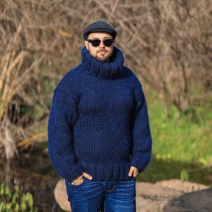 May include: A man wearing a dark blue turtleneck sweater, a dark blue newsboy cap, and dark sunglasses. He is standing in front of a background of brown and green foliage.