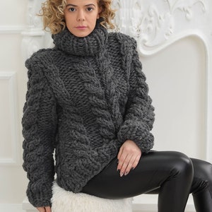 May include: A woman wearing a grey chunky knit turtleneck sweater and black leather trousers sits on a white furry stool. She is wearing black high-top trainers with white soles.