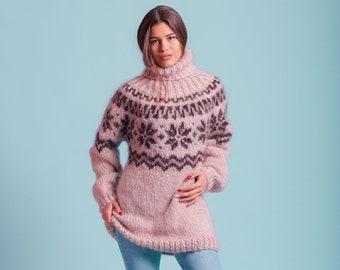 May include: A woman wearing a pink and black patterned sweater with a turtleneck and blue jeans. She is standing on a light blue background.